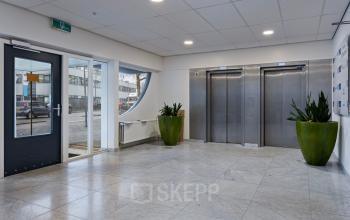 De entree van het kantoorpand aan de Brauwweg 52-66 in Schiedam. Moderne stenen vloer, groene planten naast liften, en ruime toegangsdeur benadrukken de functionele designstijl.