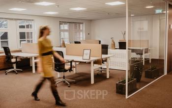 Moderne kantoorruimte met glazen wanden en bureaus op Bredaseweg 8, Breda Terheijden. Persoon in gele jurk loopt door de ruimte, geschikt voor kantoren of huren.