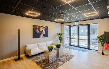 Moderne ontvangstruimte in een kantoorpand met een stijlvolle witte bank, glazen salontafel en kunst aan de muur. Gezellige verlichting en groene planten verfraaien de ruimte. Ideaal voor kantoorruimte huren.