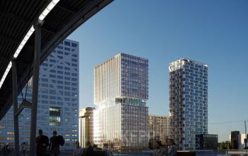 Moderne kantoorcomplexen bij Utrecht Centraal Station, gelegen aan Stadsplateau 3, met glanzende glazen buitenkant reflecterend in het zonlicht. Mensen zitten op bankjes en wandelen in een rustgevende omgeving.