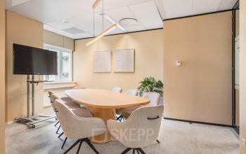 Vergaderruimte in kantoorpand te Utrecht Centraal Station met ovale houten tafel, acht stoelen, en een groot scherm. Neutrale kleuren en natuurlijke verlichting geven een zakelijke sfeer.