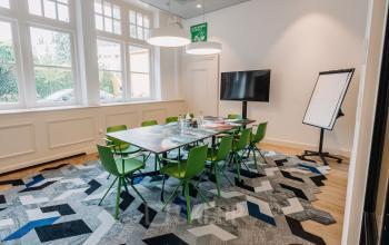 Moderne vergaderruimte in kantoorpand Kromme Nieuwegracht 3, Utrecht Centrum met kleurrijk tapijt, grote tafel, groene stoelen, en presentatiescherm.