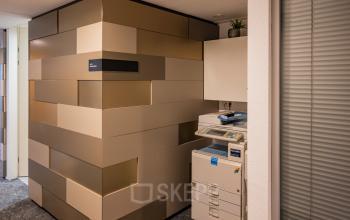 Binnen een kantoorpand op Middenburcht 136, Utrecht Leidsche Rijn staat een kopieerapparaat naast een modern ontworpen muur. Ideale plek voor bedrijven om hun dagelijkse taken efficiënt uit te voeren.