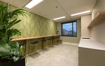 Stijlvolle kantoorruimte huren aan de Varrolaan in Utrecht Rijnsweerd, met hoge krukken bij een lange tafel en een groene plantenbak, perfect voor informele bijeenkomsten of pauzes met uitzicht.