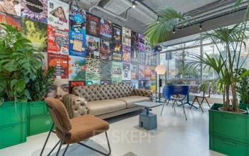 Gezellige werkruimte met kleurrijke posters, geschikt voor kantoorruimte huren in Utrecht Rijnsweerd. Voorzien van een comfortabele bank, stoelen, planten en grote ramen voor natuurlijk lichtinval.
