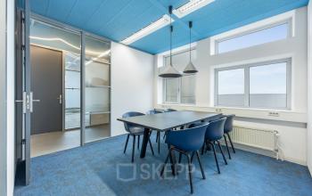 Moderne, goed verlichte kantoorruimte met blauwe accenten, voorzien van een tafel en zes stoelen, ideaal voor vergaderingen of als werkruimte. Grote ramen bieden veel natuurlijk lichtinzichten. Geschikt voor kantoorpand huren.