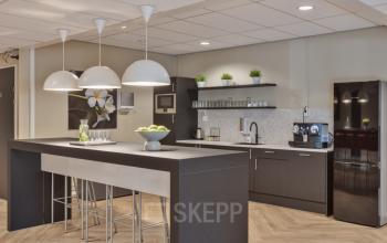 Stijlvolle pantry met een lange tafel, moderne verlichting, een koelkast en koffiezetapparaat in een kantoorpand op Niasstraat 1, Utrecht West. Ideaal voor pauzes of informele meetings.