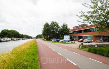 Exterieur van het kantoorpand aan Kanaalweg 22 in Utrecht Zuid, met een moderne rode bakstenen gevel en parkeerplaats. Groenrijke omgeving langs het kanaal met auto’s op de achtergrond.