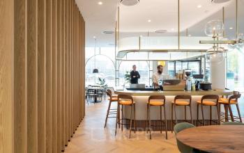 Stijlvolle kantine in het kantoorpand aan Europalaan 40, Utrecht Zuid, met houten meubels, moderne verlichting en twee mensen die koffie bereiden. Ideale werkruimte huren met een gezellige sfeer.