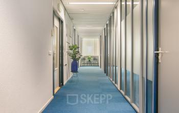 Lange gang in een kantoorpand met blauwe vloerbedekking en glazen wanden aan Vendelier 61, Veenendaal. Rechts zijn meerdere deuren zichtbaar, met een tafel en plant aan het einde van de gang. Ideaal voor kantoorruimte huren.