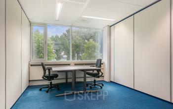 Kantoorruimte huren in Veenendaal met natuurlijk licht, neutrale muren en blauwe vloerbedekking. Gelegen op Vendelier 61, ideaal voor vergaderingen en werk. Ruimte is gemeubileerd met stoelen en een tafel.
