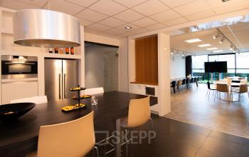 Een moderne kantoorruimte op Gildestraat 13, Velp met een gemeubileerde open vloer ruimte, houten tafel en aanrecht. Ideaal voor kantoorruimte huren. Op de achtergrond is een grotere vergadertafel zichtbaar.