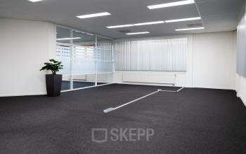 Kantoorruimte huren in het kantoorpand aan Parlevinkerweg 1, Venlo. Modern interieur met veel daglicht en grijze vloerbedekking. Inclusief raam met jaloezieën en een groene plant in een hoek.