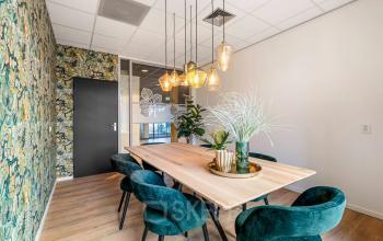 Kantoorruimte huren aan Agro Business Park 22, Wageningen. Gezellige vergaderruimte met houten tafel, stijlvolle stoelen, en opvallende wanddecoratie. Hanglampen creëren een warme sfeer.