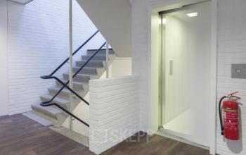 Ingang van een kantoorpand op Pompmolenlaan 9, Woerden, met een open lift en trap. Ideaal voor wie kantoorruimte wil huren. Heldere verlichting en praktische indeling creëren een professionele uitstraling.