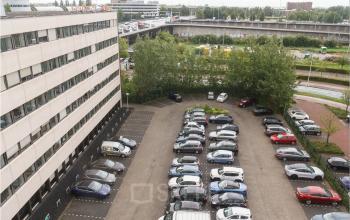Parkeerterrein naast kantoorpand Bredewater 26 in Zoetermeer, met meerdere geparkeerde auto's en omringende groene bomen.