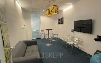 Kantoorruimte huren aan de Baron de Coubertinlaan 35 in Zoetermeer met een grijze bank, twee stoelen, hoge tafel en een tv aan de muur.