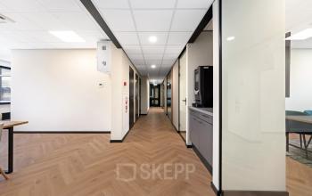 Ruime gang in een kantoorpand met lichte muren, glazen zijwanden, en houten vloer. Aan de linkerzijde staat een koffiezetapparaat. Ideaal voor bedrijven die een kantoorruimte willen huren op Stationsplein 4 in Zwijndrecht.