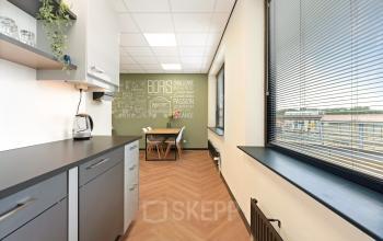 Kantoorruimte huren op Stationsplein 4, Zwijndrecht. Moderne pantry met lichtinval via grote ramen, uitgerust met keukenkastjes, beklede vloer en stoelen. Ideaal voor zakelijke pauzes en informele vergaderingen.