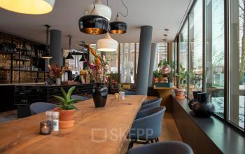 Lichte en smaakvolle kantoorruimte met lange houten tafel, moderne verlichting en veel groen. Ideaal voor vergaderingen of informeel overleg.