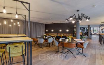 Moderne kantoorruimte met houten vloeren en stijlvolle verlichting, tafels met stoelen en hoge zitplekken. Gelegen op Assendorperdijk 1, Zwolle. Geschikt voor kantoorruimte huren.