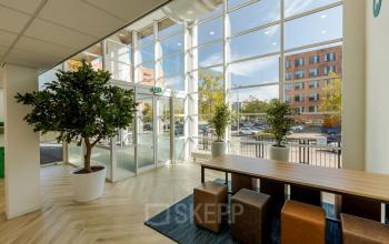 Lichte entree van een kantoorpand op Zuiderzeelaan 15-25 in Zwolle, met grote ramen, houten tafel en krukken; ideaal voor mensen die kantoorruimte willen huren.